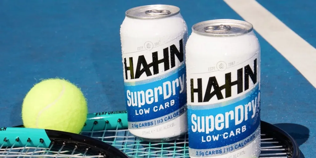 Close-up photo of two frosty cans of Hahn SuperDry sitting on a tennis racket next to a tennis ball