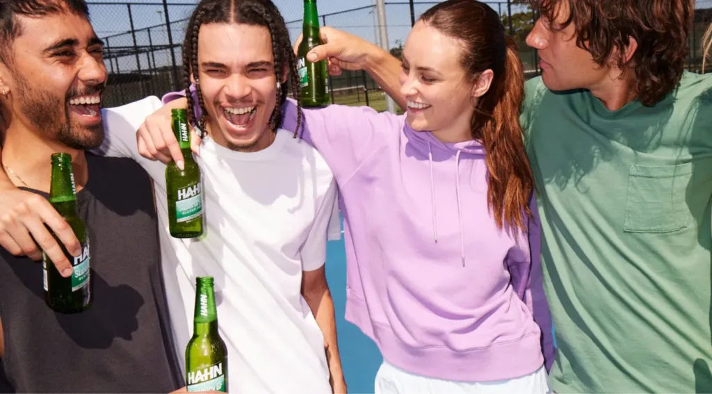 Four friends smiling while holding Hahn SuperDry Gluten Free bottles on a sports court